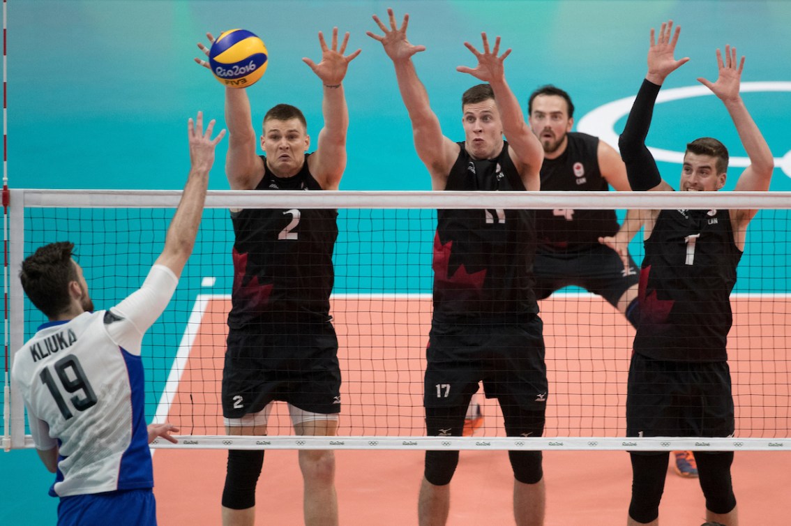 Canadian volleyball players in black prepare to block the ball hit by their opponent 