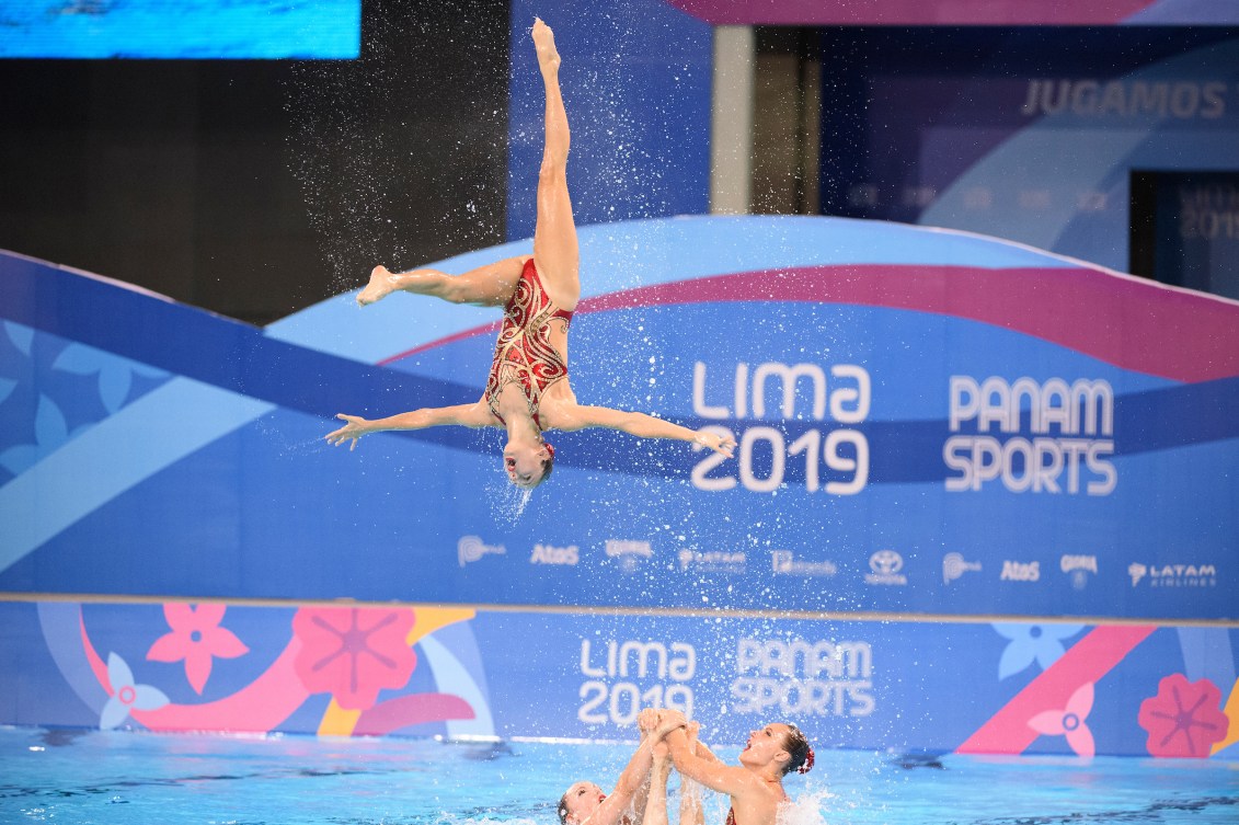 An artistic swimming does a backflip in the air abover her teammates
