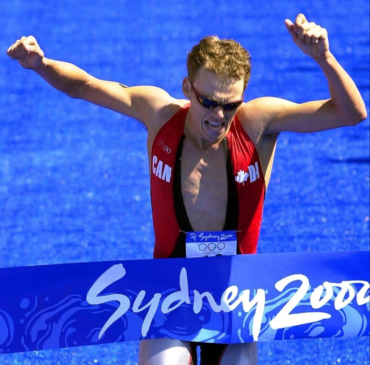 Simon Whitfield raises his arms as he runs into a blue banner at the finish line 