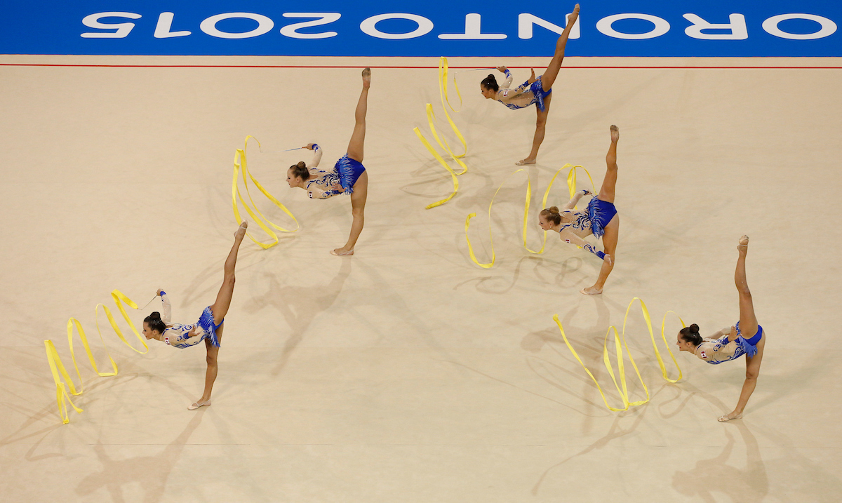 Five rhythmic gymnasts stand in a split with one leg above head while swirling their ribbons 