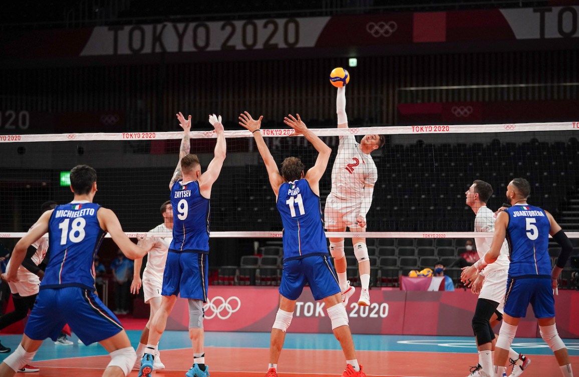 Canadian volleyball player in white hits the ball over the net towards opponents in blue 