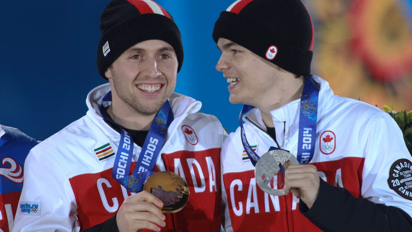 Mikael Kingsbury looks at Alex Bilodeau laughing who is standing at the Sochi 2014 medal presentation. Alex Bilodeau is smiling and holding his bronze medal and Kingsbury is holding him silver medal. They are dressed in their red and white Team Canada sweaters and toques.
