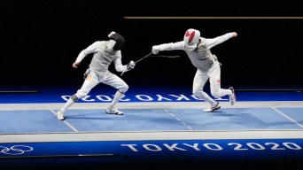 Two fencers swordfight on a blue piste in a dark arena