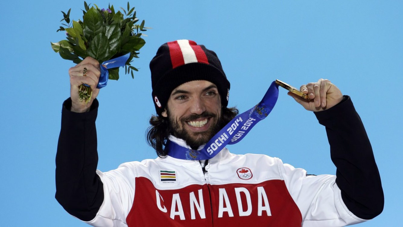 Charles Hamelin on the podium at Sochi, holding up flowers in his right hand and his medal in the other.