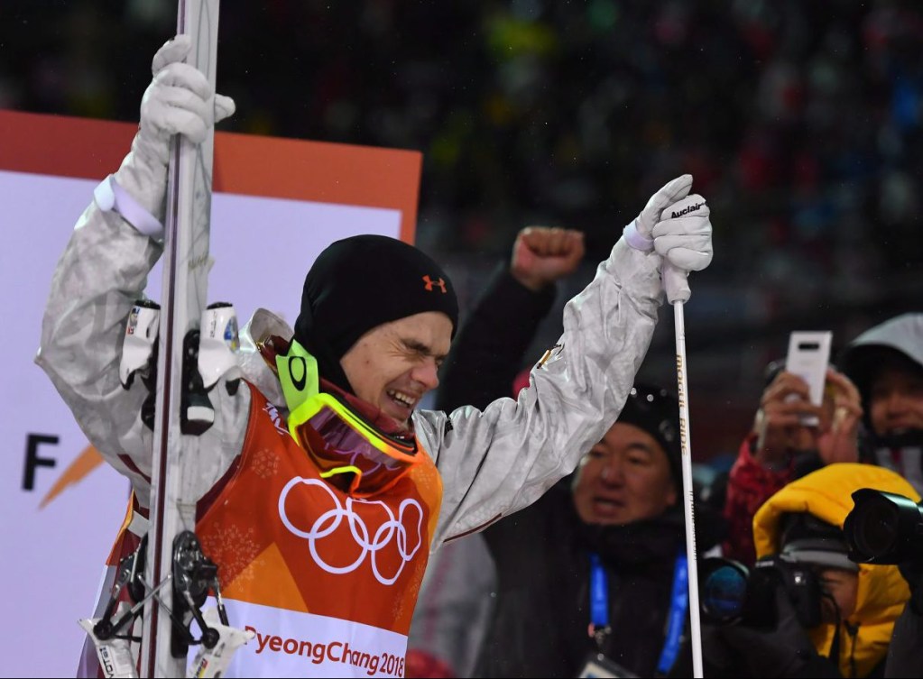 Team Canada skier Mikael Kingsbury holds his skis and poles above his head cheering after winning his first Olympic title in the men's moguls at the 2018 Winter Olympic Games in Pyeongchang. His eyes are closed and he is smiling widely. The audience cheers and takes photos in the background.
