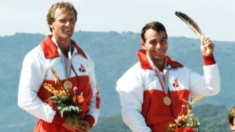 Alwyn Morris holds an eagle feather above his head while standing next to Hugh Fisher on the podium