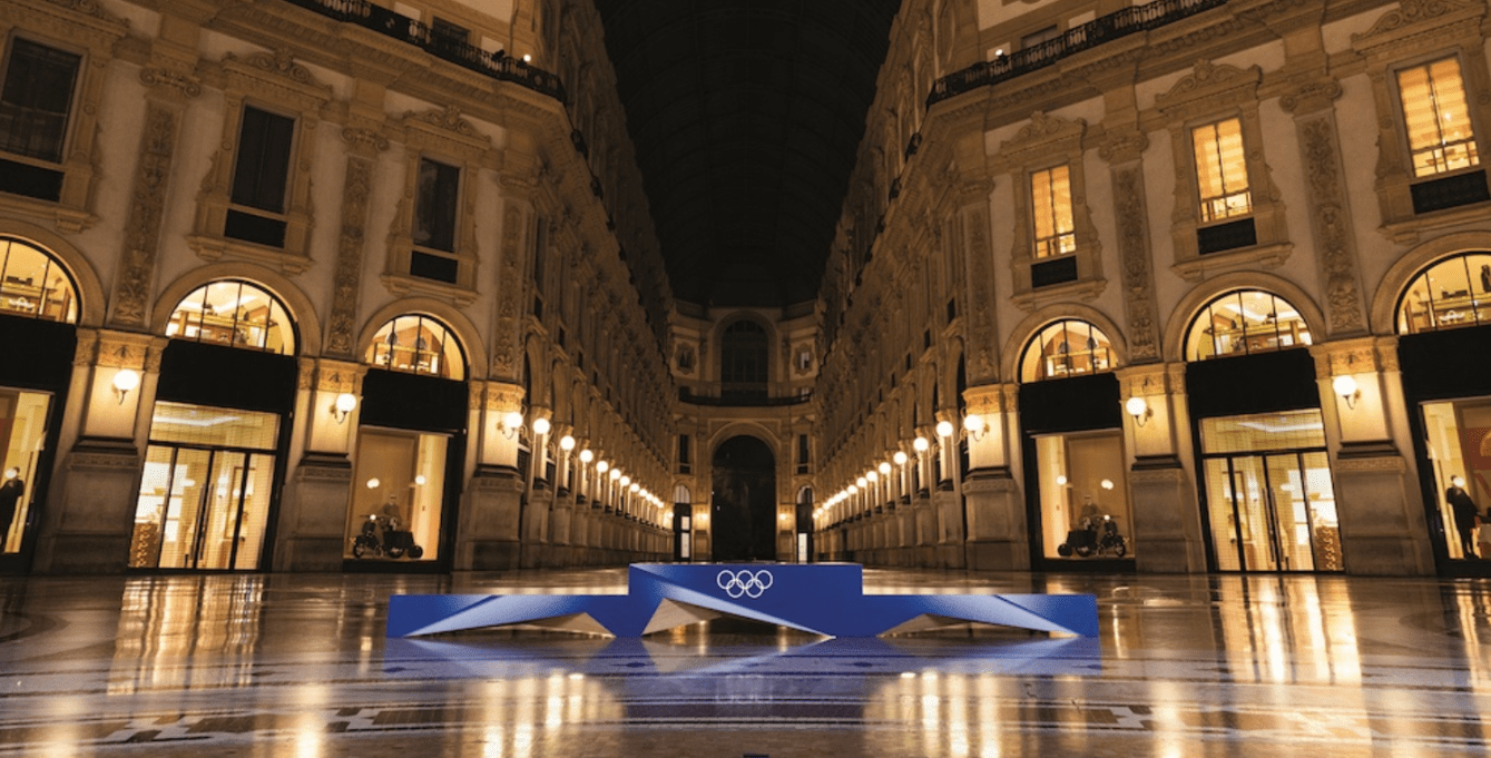A shot of the blue Milano Cortina 2026 podium in front of a brightly lit town square at night