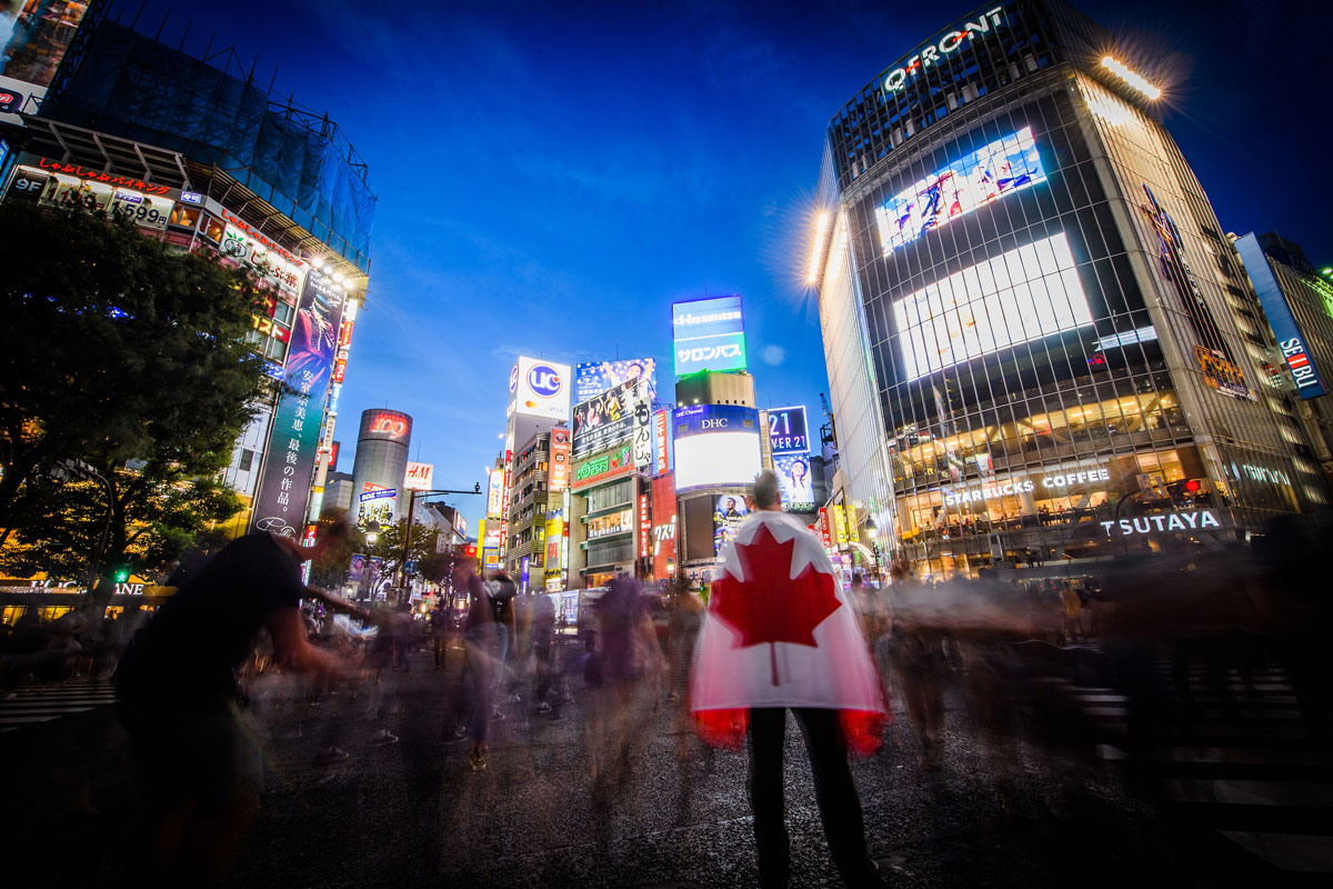 A man holding a Team Canada flag stares out at the crowds walking through Shibuya Crossing.
