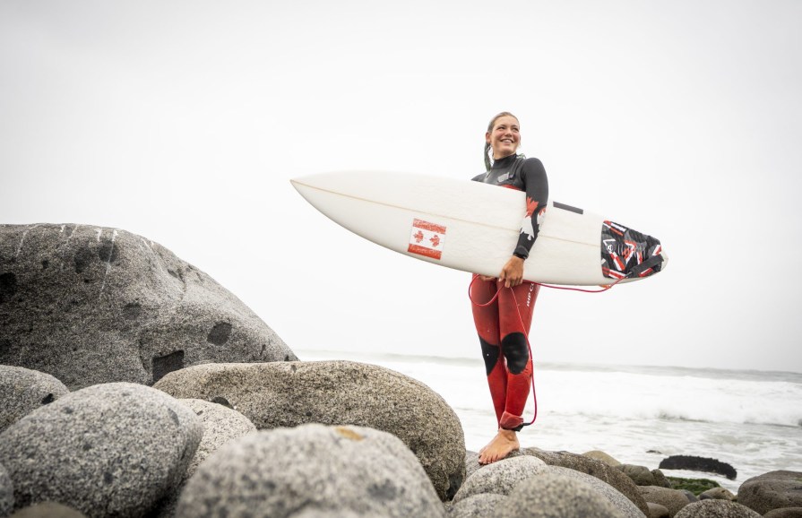 team-canada-lima-surfing woman stands on rocks with a surf board