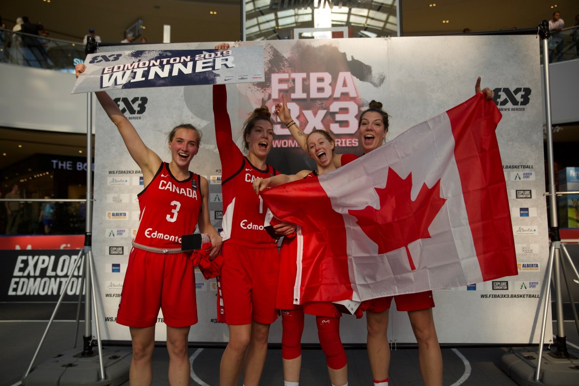 Canada's 3x3 team celebrates their victory with Paige (far left) and Michelle holding a '3x3 Edmonton 2019 Winner' sign, and Catherine and Katherine (far right) holding the Canadian flag.