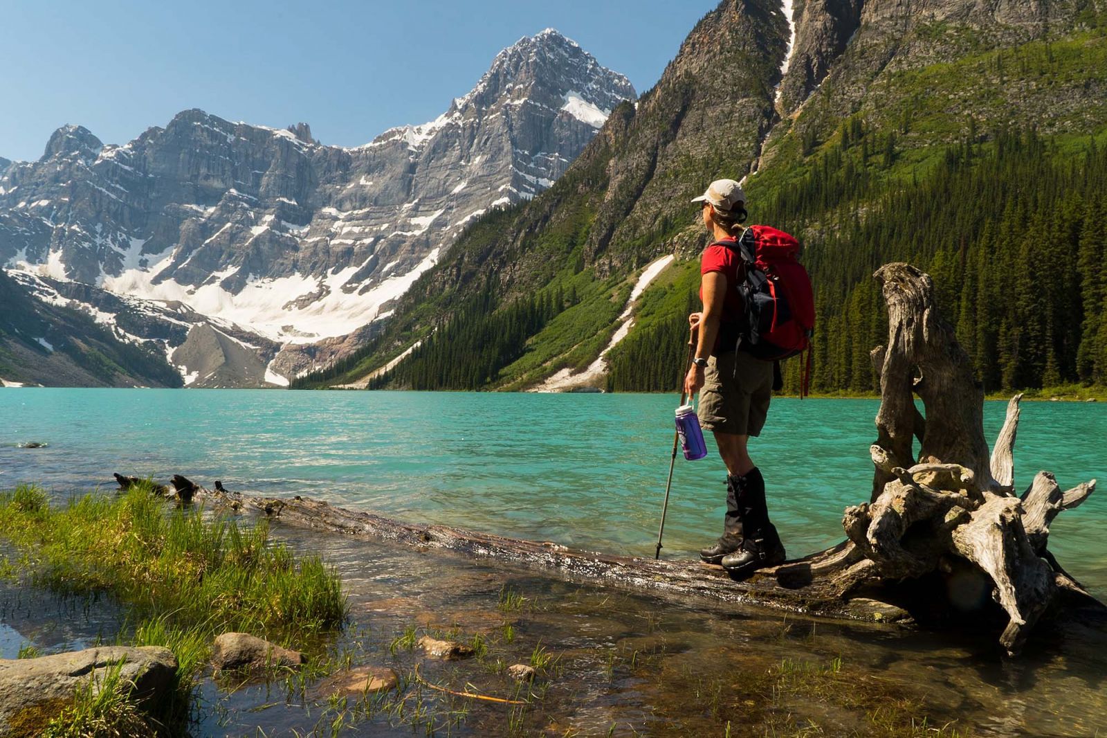 A hiker stands beside the turquoise water and looks out at the Mountains.