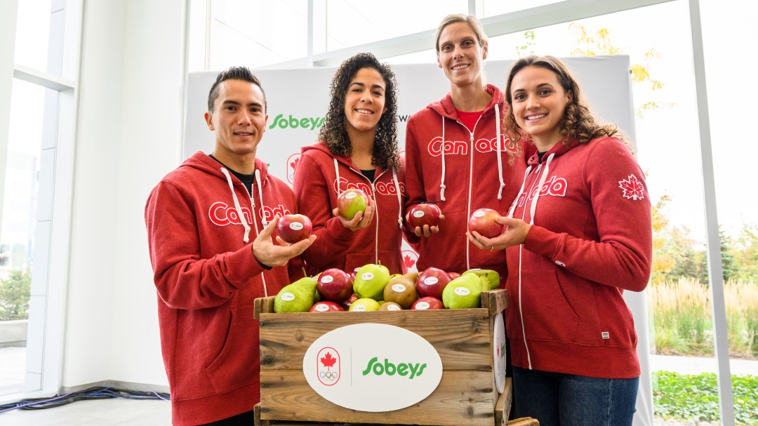 (left to right) Sean McColl, Kia Nurse, Sarah Pavan and Kylie Masse each hold an apple and stand behind a carton of apples with a Team Canada and Sobeys logo lock.