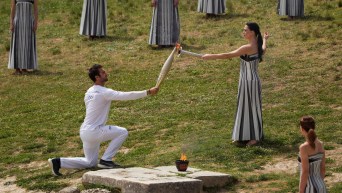 A woman dressed in an ancient Greek dress holds a flamed torch to light the Olympic torch held by a man dressed in a white athletic uniform