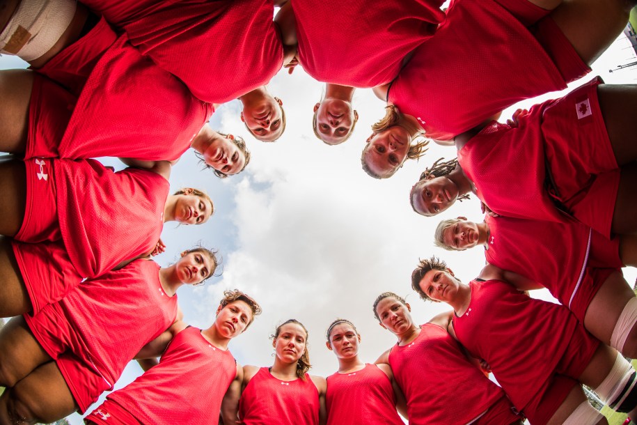 A shot from the ground up of the women's rugby team looks down at the ground while standing in a circle.