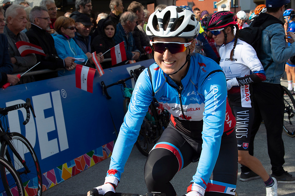 Leah Kirchmann at a competition. She is in the foreground of the picture with fans and other cyclists in the background. She is wearing a white helmet, a blue and white shirt, and black bike shorts with red and blue detailing.