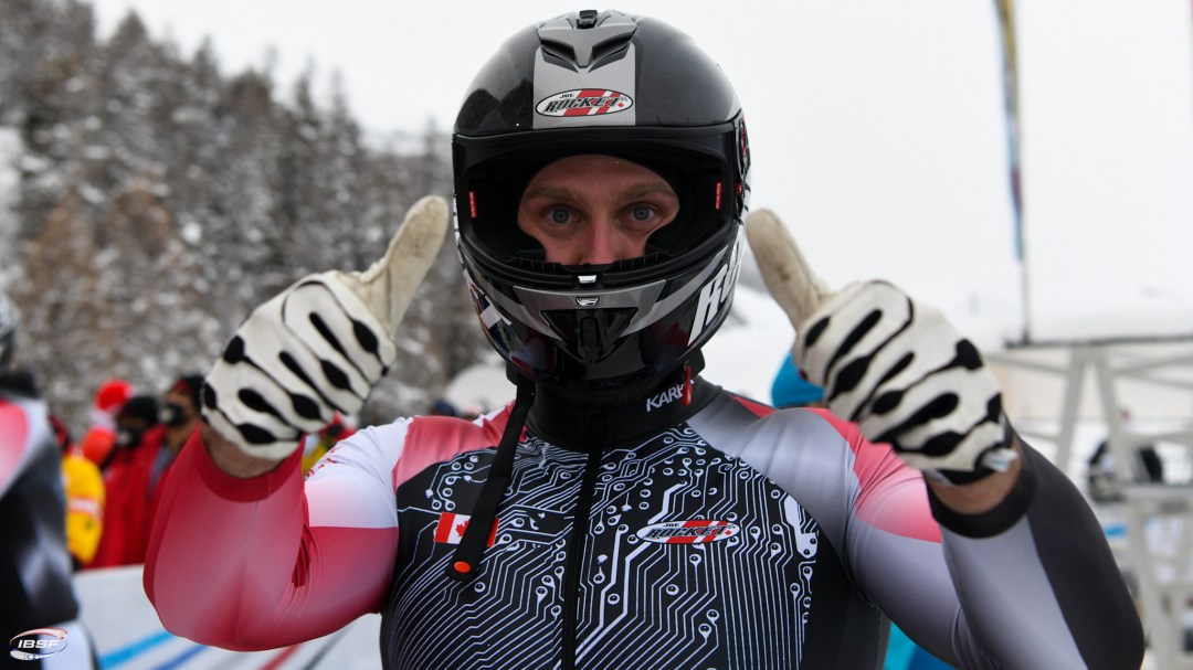 A male bobsledder gives a thumbs up with both hands. He is wearing his helmet but the visor isn't on so we can see his eyes.