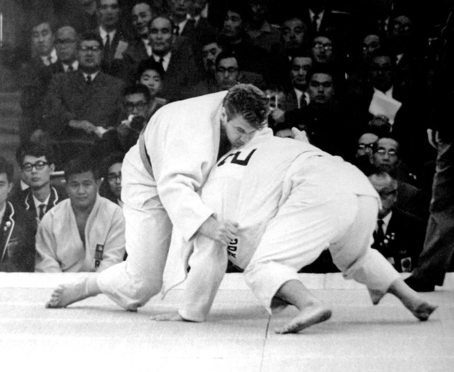 In a black and white photo, Doug Rogers holds his opponent on a judo mat in front of spectators at the 1964 Olympics.