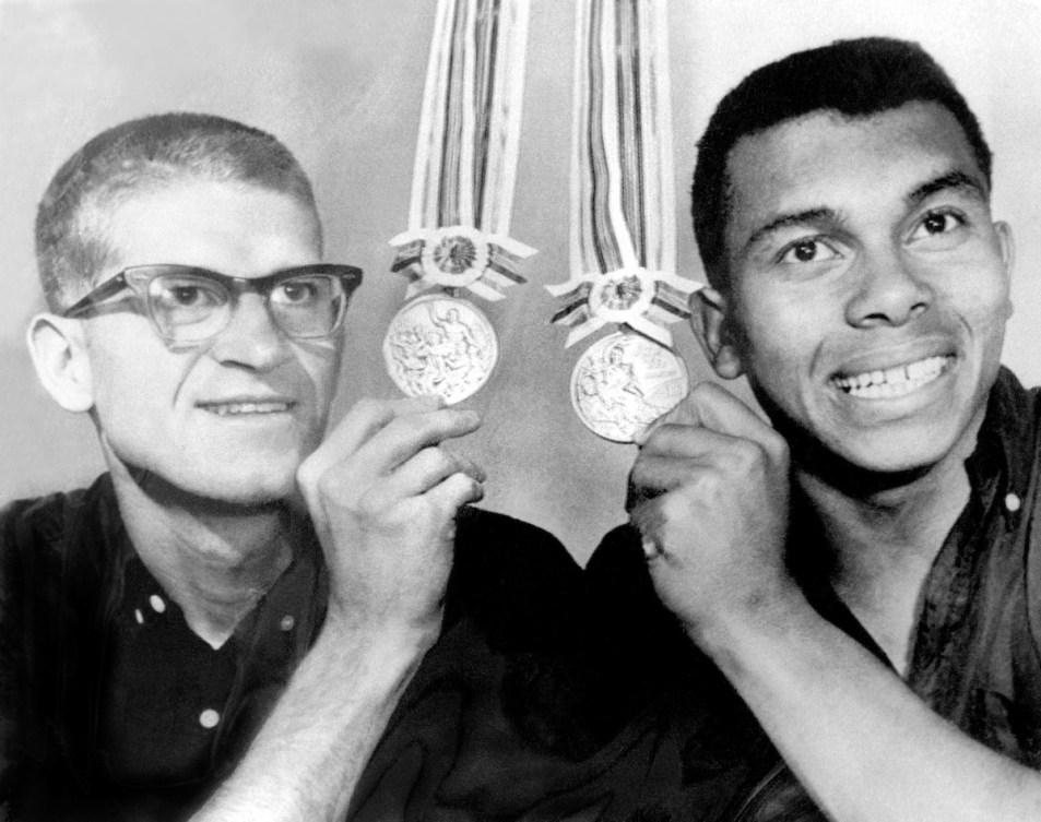 In a black and white photo, a smiling Bill Crothers and Harry Jerome display their medals won at the 1964 Olympics.