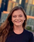 Head shot of Amy Stoparczyk. She has long brown hair and is wearing a black shirt. The city skyline is behind her, out-of-focus.