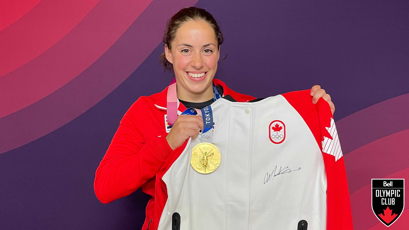 woman stands in front of a red background holding a gold medal and a white jacket with a COC logo, Hudson's Bay logo and a black signature on it