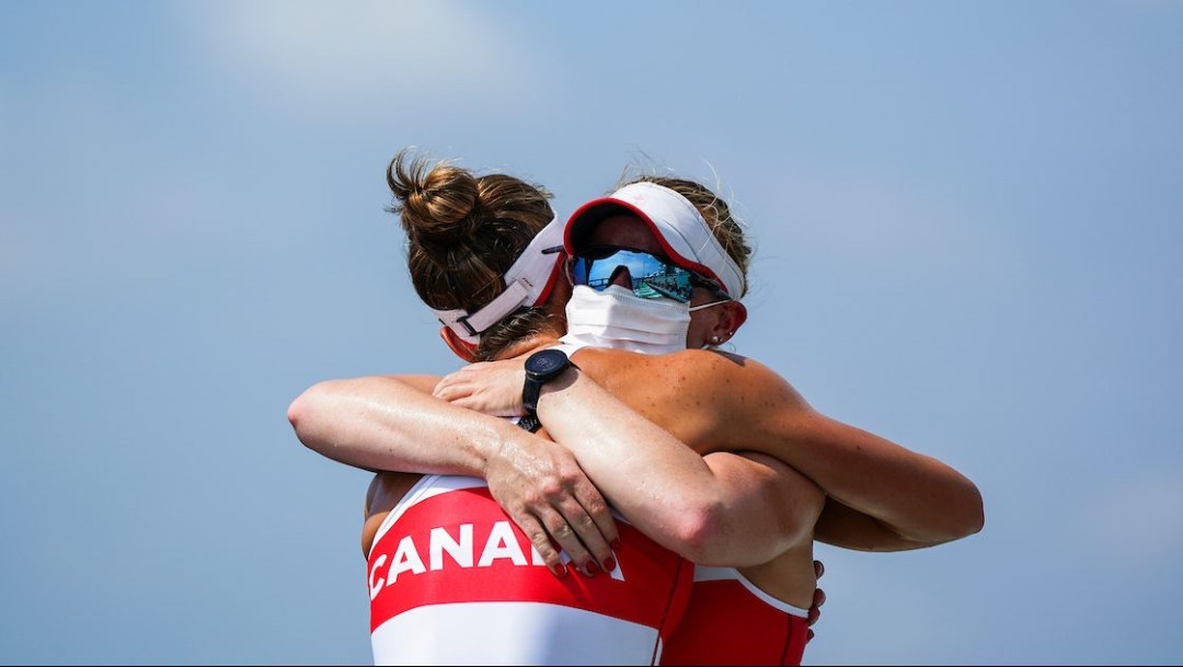 Rowers Caileigh Filmer and Hillary Jenssens, wearing sunglasses and protective masks, hug in celebration against the backdrop of a clear blue sky.