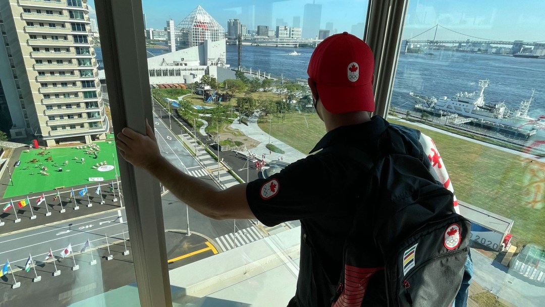 A man wearing a Team Canada baseball cap backwards looks out at the view of Tokyo Bay.