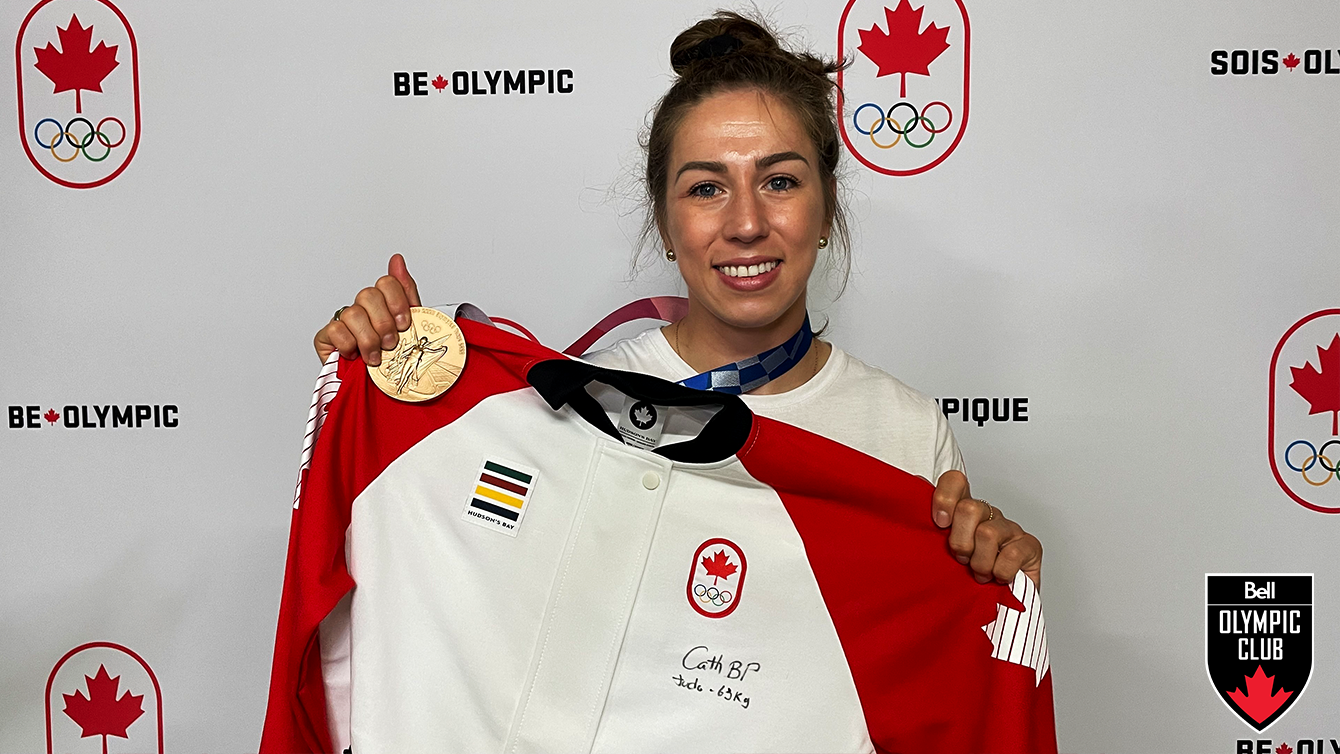 catherine stands in front of white backdrop holding a red and white jacket and a bronze olympic medal