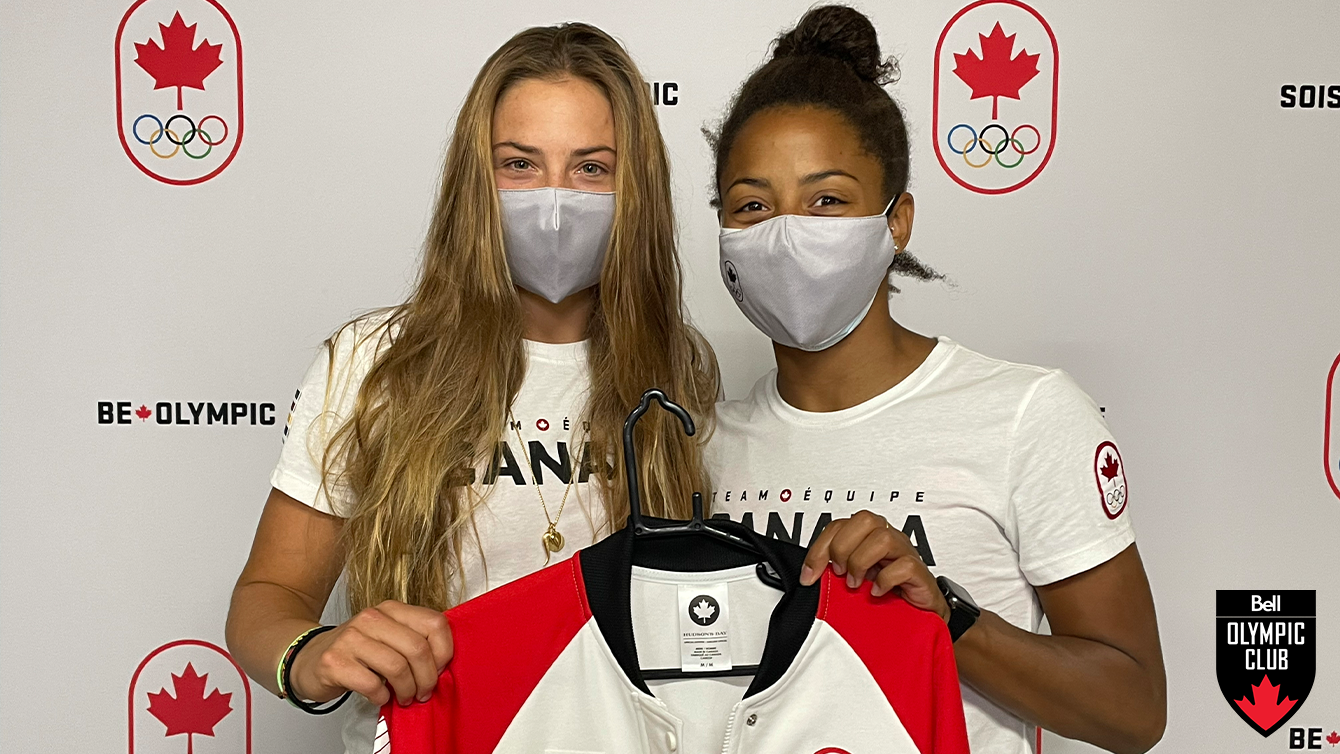 two women stand in front of white background holding a white jacket that has a COC logo, hudsons bay logo and signature on it