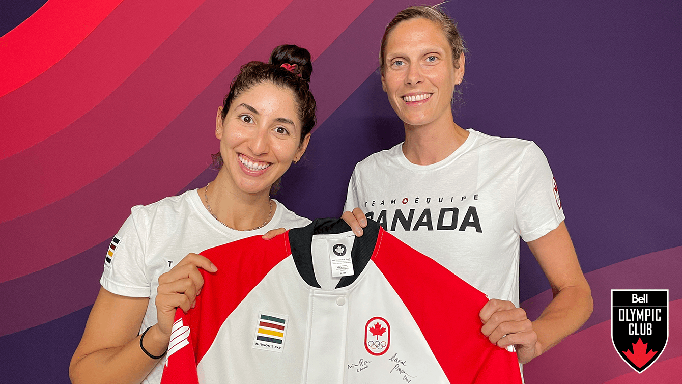 two women stand in front of red background holding a white jacket that has a COC logo, hudsons bay logo and signature on it