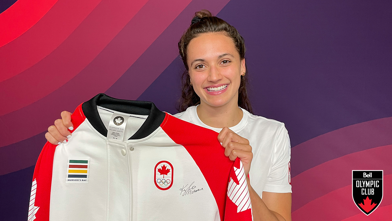 woman stands in front of red background holding a white jacket that has a COC logo, hudsons bay logo and signature on it