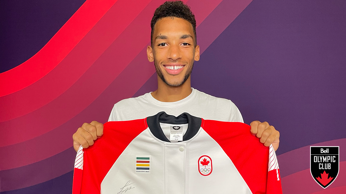 athlete stands in front of a red background holding a white jacket with a team canada logo, Hudsons' Bay logo and signature on it.