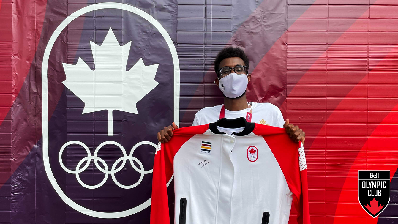 man stands in front of a wall with the COC logo (white maple leaf above olympic rings graphic) holding a white jacket with a signature on it