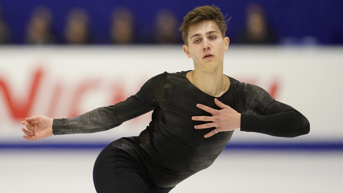 Roman Sadovsky étend le bras droit et touche son coeur de la main gauche tout en regardant la caméra lors d'une compétition de patinage artistique