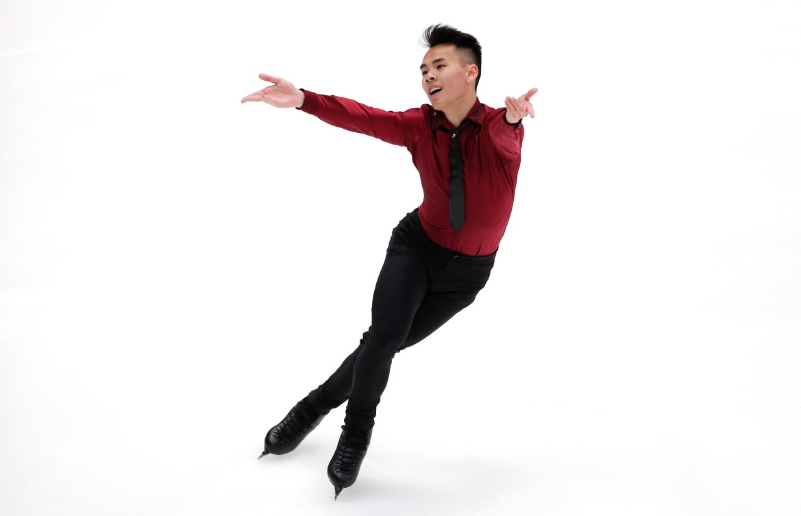 Nam performing a routine. His legs are crossed over and he is holding his arms out. He is wearing a dark red long sleeved button up shirt and a black tie, along with black pants and black skates.