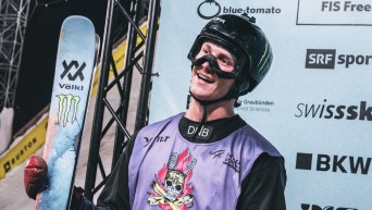 Teal Harle smiles while waiting for his results. He is wearing his goggles and helmet, while holding his skis vertically.