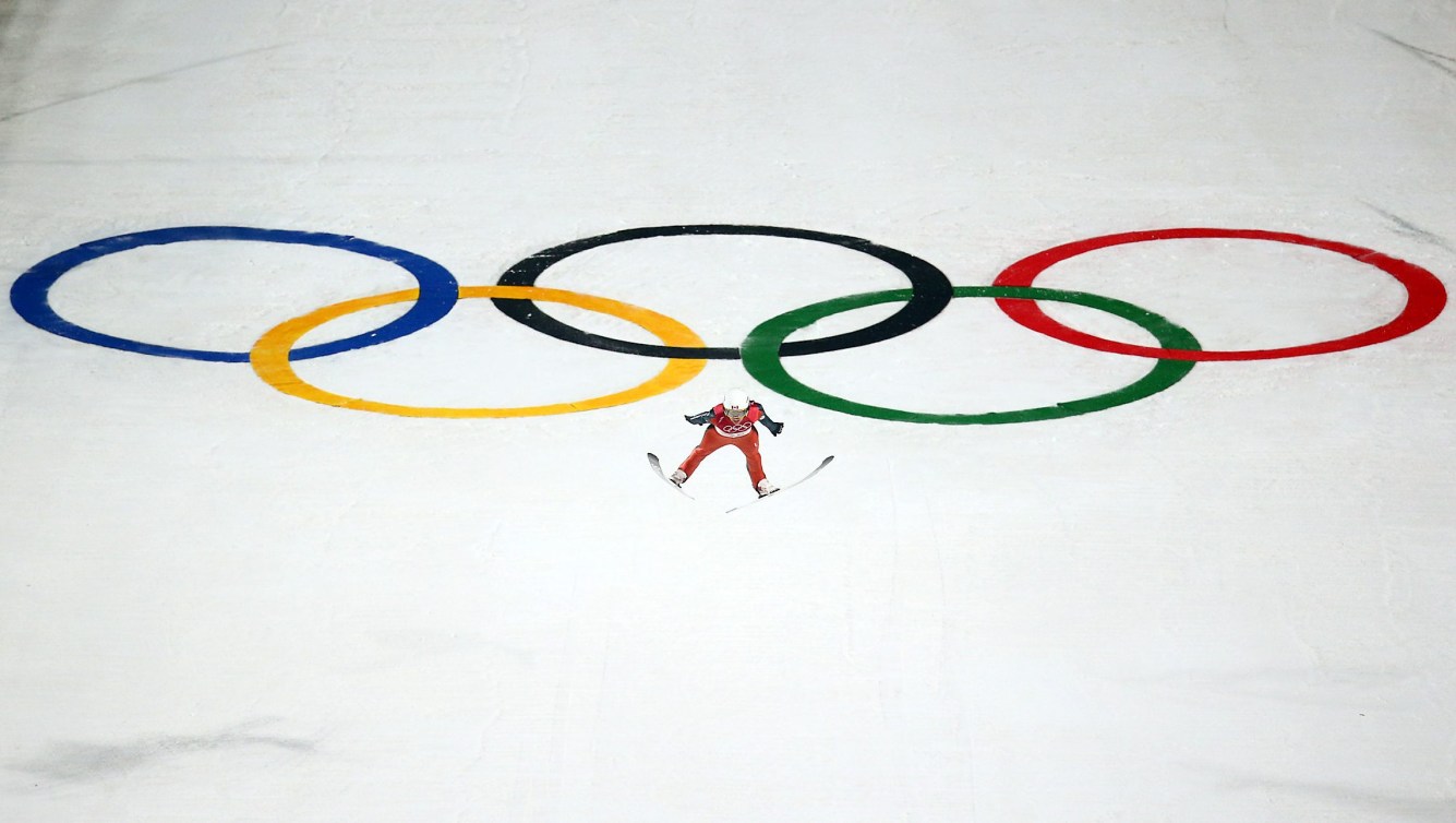 Ski jumper flies through air with Olympic rings in background on the snow