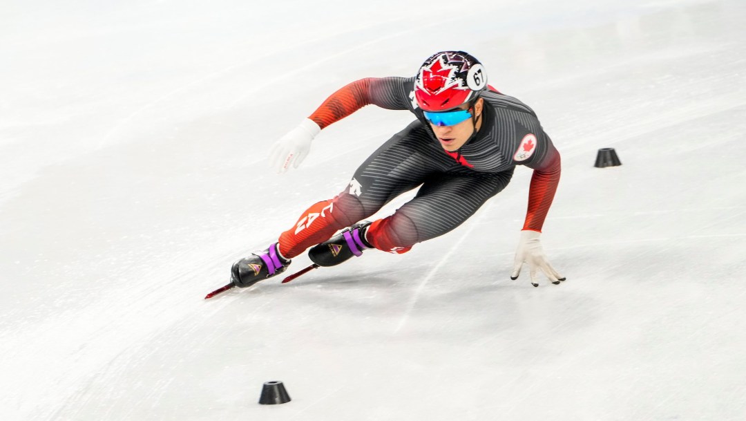 Short track speed skater in red and black suit makes a turn around little black pylons