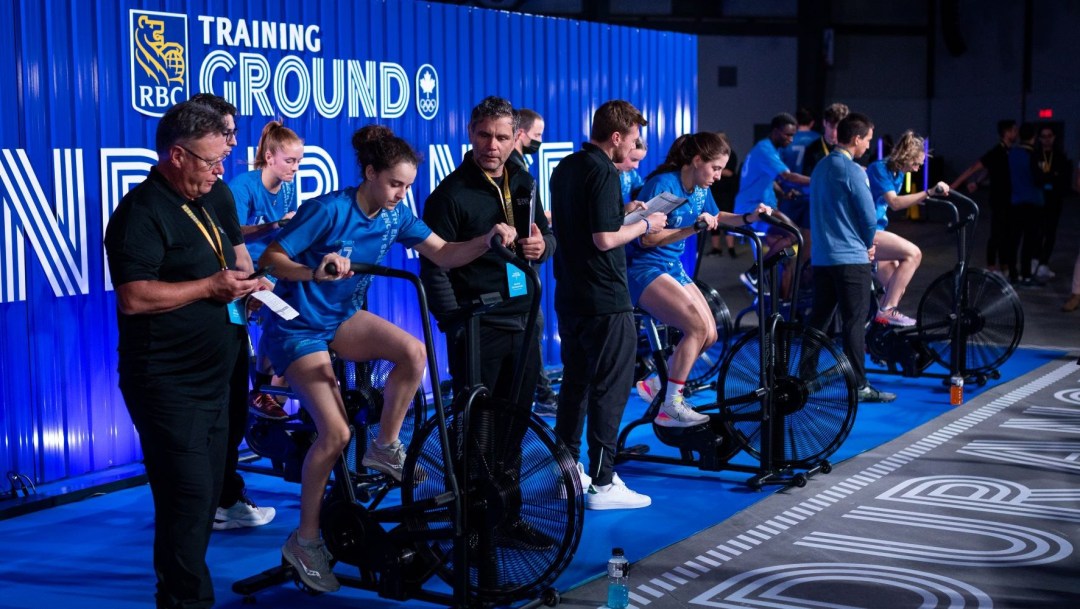 Four athletes in blue RBC t shirts ride stationary bikes under the watchful eye of recruiters