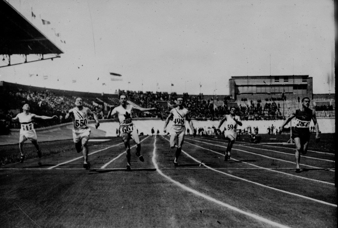Black and white photo of men racing across a finish line in a sprint event 