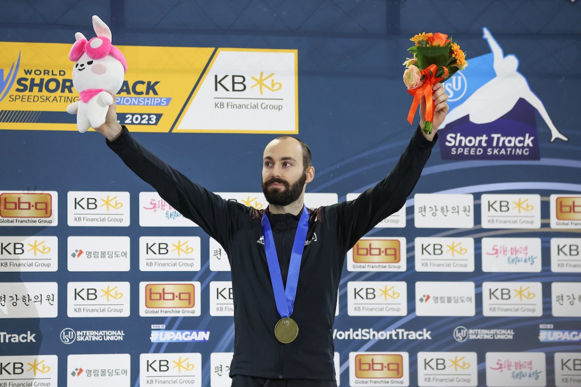 Steven Dubois wears bronze medal on podium while holding up flowers and mascot toy 