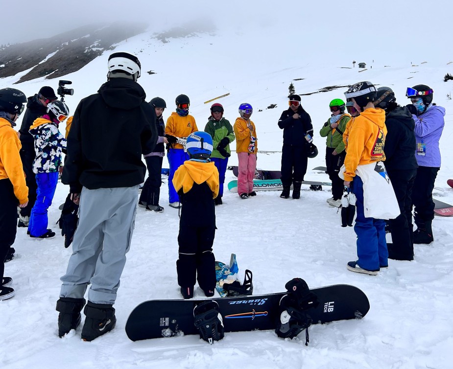 A group of young Indigenous snowboarders stand in a circle for a land acknowledgement and blessing