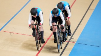 Three female track cyclists in blue jerseys form a line on the velodrome track