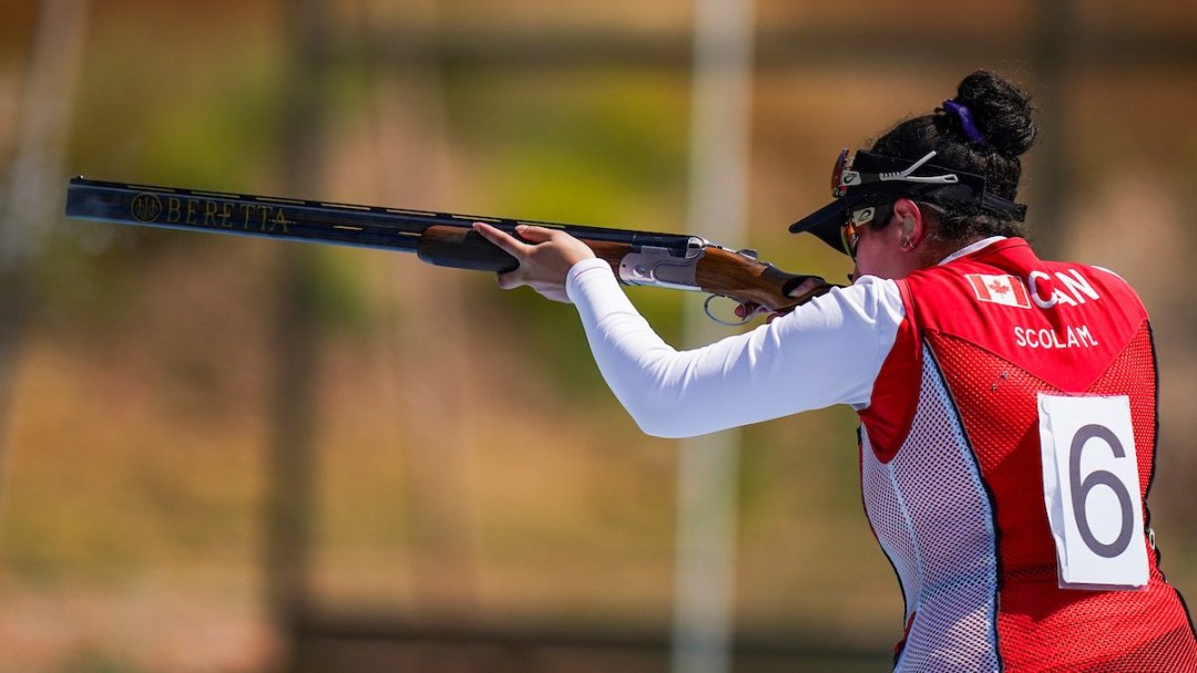 A trap shooter in a red vest holds her shotgun up prepared to shoot