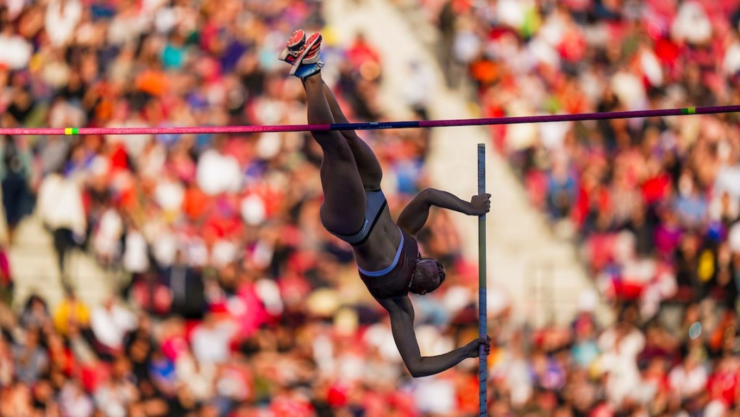 Rachel Hyink is almost over the bar while performing a pole vault, with a blurred background of the packed stadium