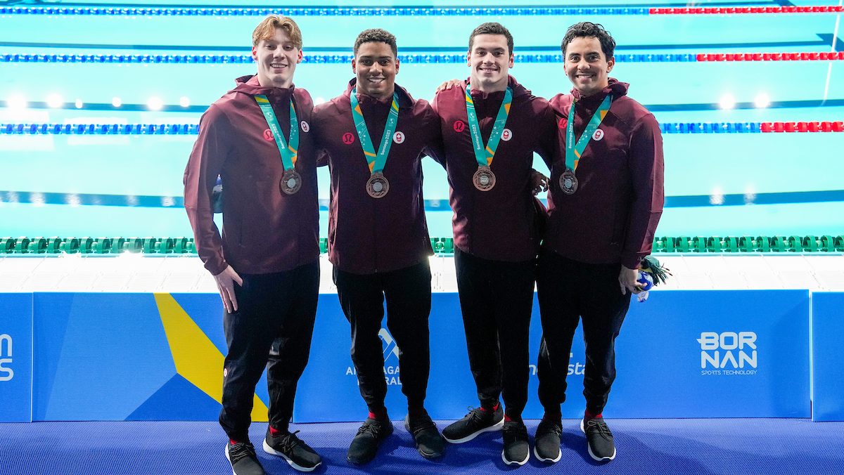 Four male swimmers stand by the pool with their bronze medals around their necks