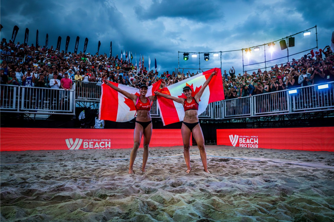 Melissa Humana-Paredes and Brandie Wilkerson pose with Canadian flags on the sand court with fans behind them