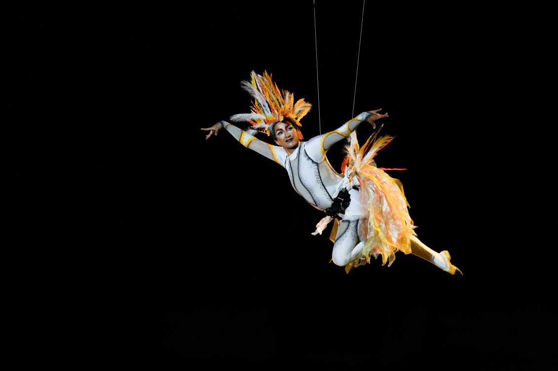 Una bailarina actúa en el aire sobre un alambre vestida como un pájaro blanco.