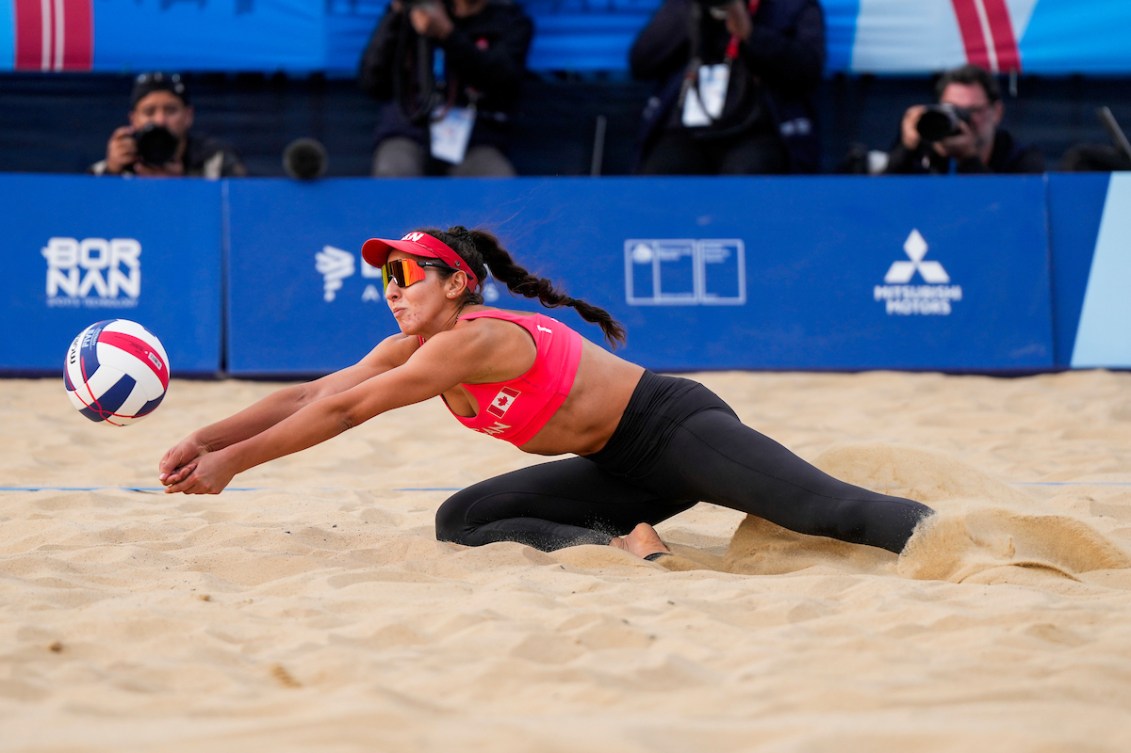 A beach volleyball player in red top and black pants kneels on the sand to bump a ball
