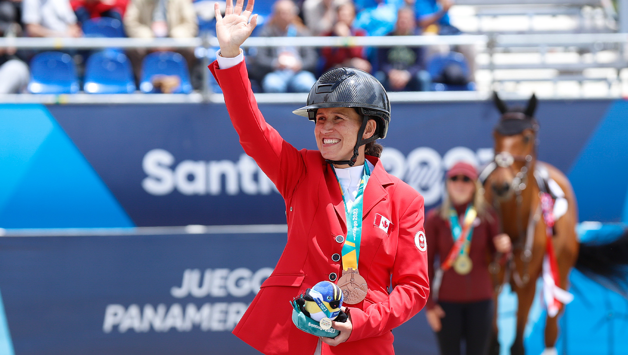 A woman wearing a black helmet and Team Canada jacket has a gold medal around her neck and waves to a crowd.