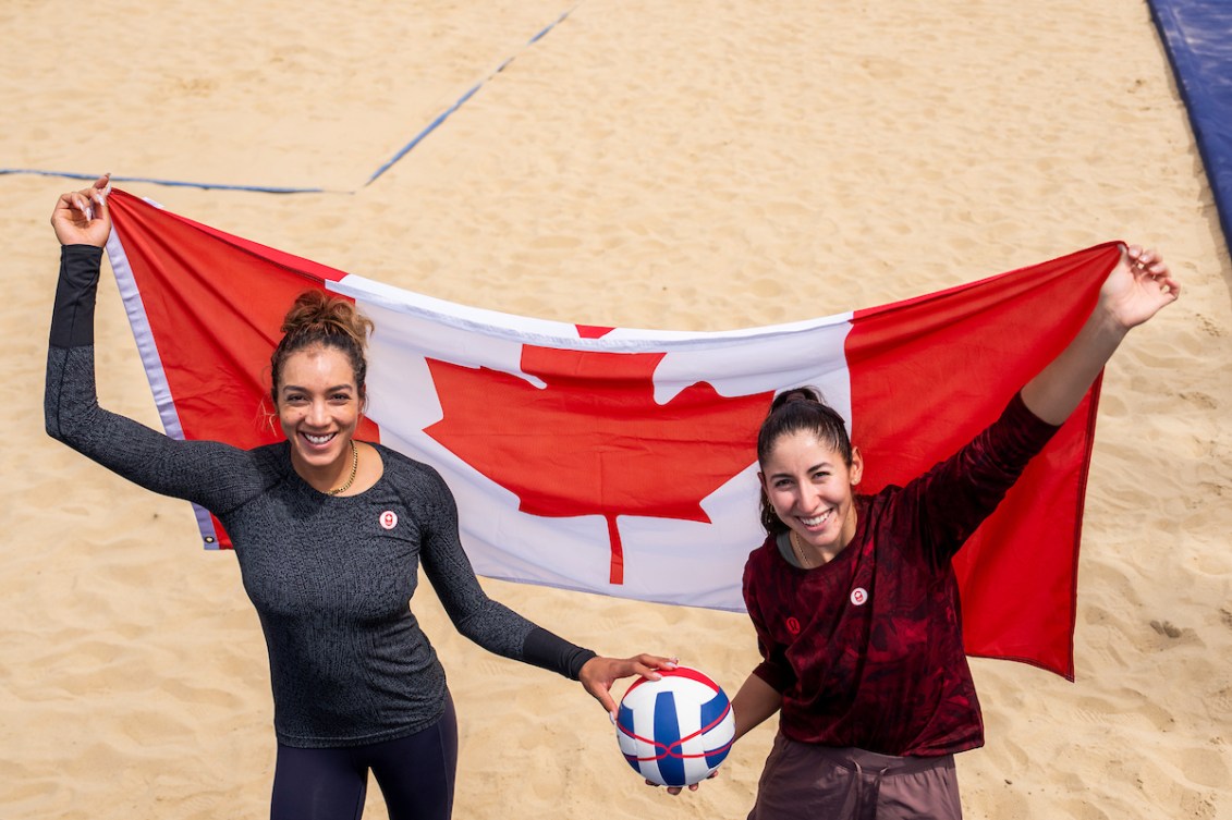 Melissa Humana-Paredes and Brandie Wilkerson pose with a Canadian flag on a beach volleyball court 